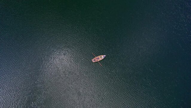 Aerial shot of a boat from above on a beautiful lake with people moving oars, the drone slowly pans up and you can see the size of the blue-green lake, edited with minimal color correction