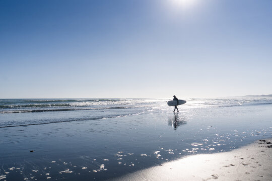 Backlit man walking along the beach with a surfboard under his arm