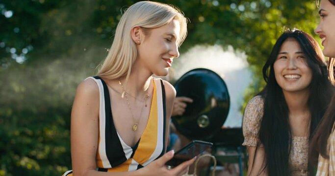 Women relax on a blanket on the grass in the open air. Friends are grilling laughing talking joking. Girls turn on favorite songs from a portable music speaker.