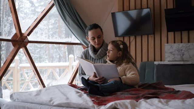 Little girl with mother reading book while sitting in bed at dome camping. Family enjoying winter glamping vacation. Scenic winter snowy nature outside