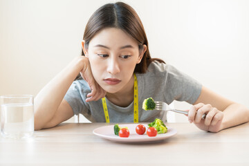 Obraz na płótnie Canvas Anorexia, unhappy beautiful asian young woman, girl on dieting, hand holding fork at broccoli in salad plate, dislike or tired with eat fresh vegetables. Nutrition of clean, healthy food good taste.