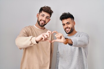 Young homosexual couple standing over white background smiling in love doing heart symbol shape...