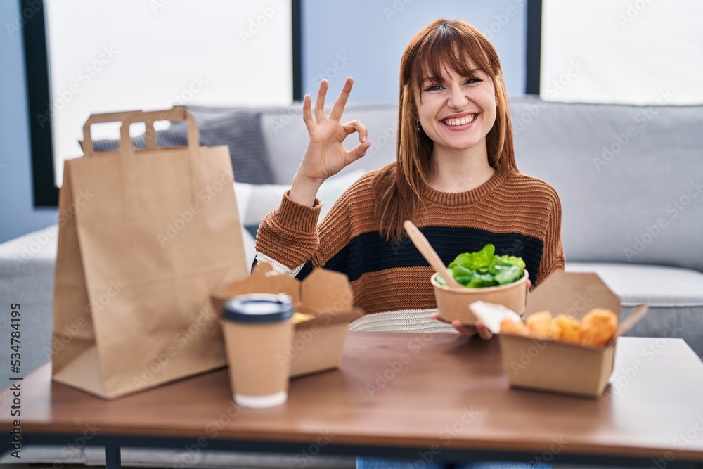 Poster Young beautiful woman eating delivery food at the living room doing ok sign with fingers, smiling friendly gesturing excellent symbol