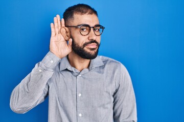 Middle east man with beard standing over blue background smiling with hand over ear listening an hearing to rumor or gossip. deafness concept.