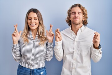 Young couple standing over blue background gesturing finger crossed smiling with hope and eyes closed. luck and superstitious concept.