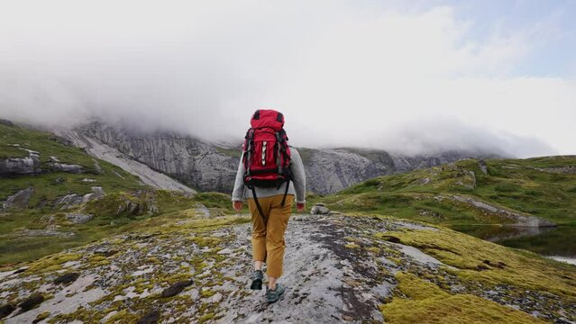 Girl with a big red backpack hikes through the highlands of Norway. The trail is barely marked. Fresh green grass. Taller mountains in the back covered partially with snow. Back view
