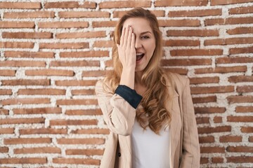 Fototapeta na wymiar Beautiful blonde woman standing over bricks wall covering one eye with hand, confident smile on face and surprise emotion.