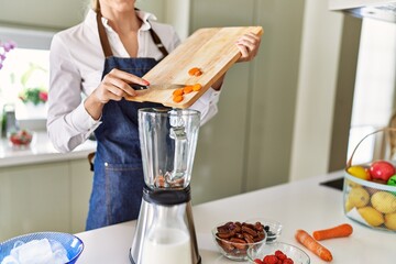 Young blonde woman pouring carrot on blender at kitchen