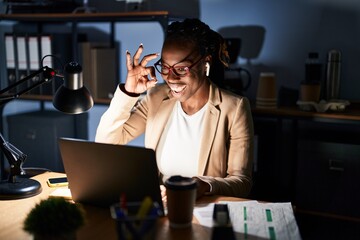Beautiful black woman working at the office at night doing ok gesture with hand smiling, eye looking through fingers with happy face.
