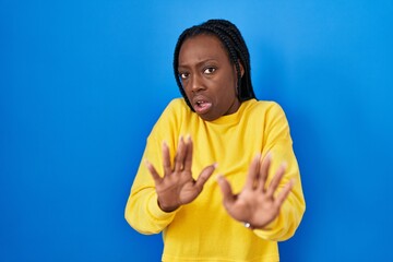 Beautiful black woman standing over blue background disgusted expression, displeased and fearful...