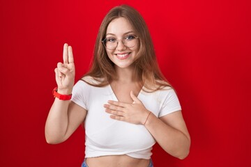 Young caucasian woman standing over red background smiling swearing with hand on chest and fingers up, making a loyalty promise oath