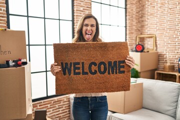 Young woman holding welcome doormat at new home sticking tongue out happy with funny expression.