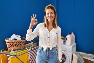 Young blonde woman at laundry room smiling positive doing ok sign with hand and fingers. successful expression.