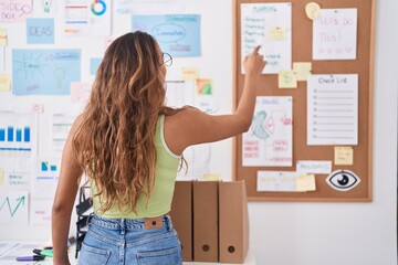 Young beautiful hispanic woman business worker looking and pointing at corkboard at office
