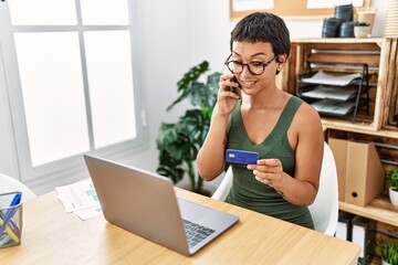 Young hispanic woman talking on the smartphone using credit card at office