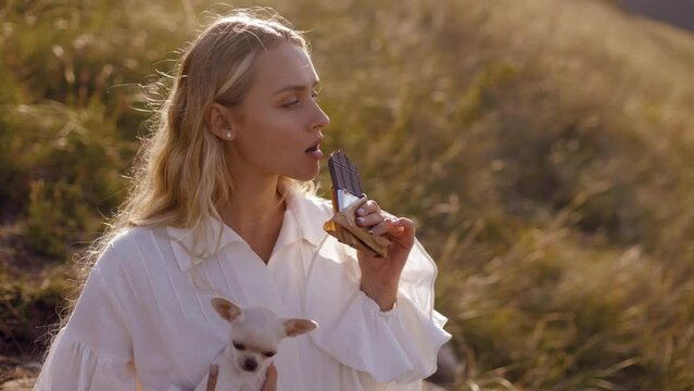 young long-haired blonde girl model with golden hair holding a white chihuahua dog sitting on a slope with an incredible view of the river biting a bar of chocolate admiring the view on a sunny autum