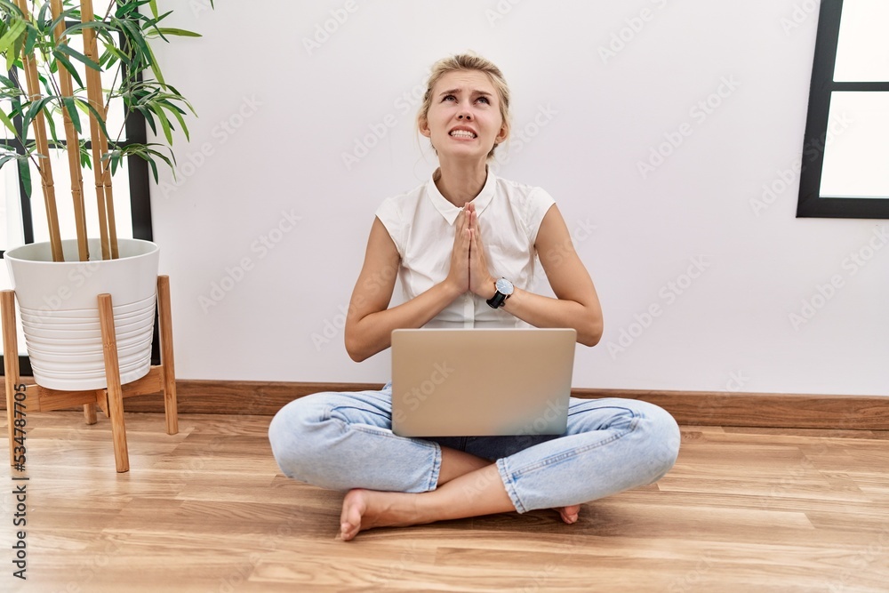 Poster Young blonde woman using computer laptop sitting on the floor at the living room begging and praying with hands together with hope expression on face very emotional and worried. begging.