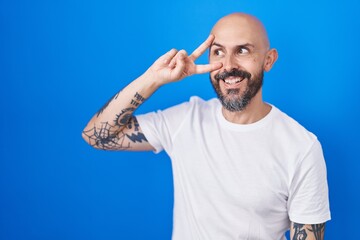 Naklejka premium Hispanic man with tattoos standing over blue background doing peace symbol with fingers over face, smiling cheerful showing victory