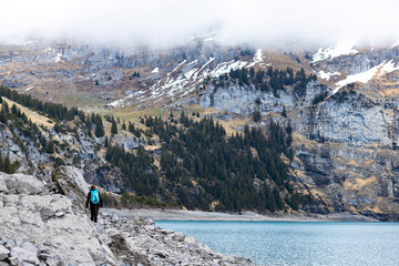 A hiker girl walks by a famous mountain lake in switzerland during gloomy weather with cloudy mountain peaks in the background; fog over Lake Oeschinensee in the swiss alps