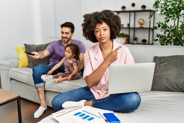 Mother of interracial family working using computer laptop at home pointing with hand finger to the side showing advertisement, serious and calm face