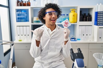 Hispanic man with curly hair working at scientist laboratory holding toxic banner pointing thumb up to the side smiling happy with open mouth