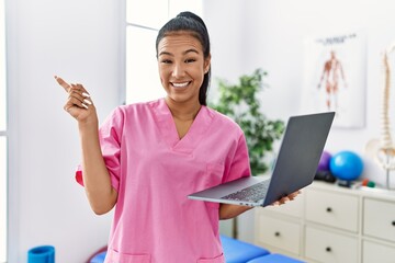 Young hispanic physiotherapist woman using computer laptop at medical clinic smiling happy pointing with hand and finger to the side