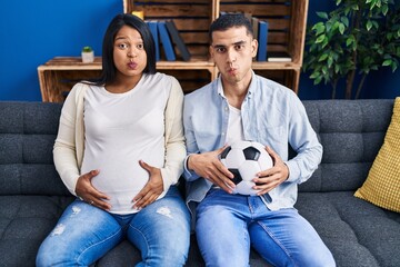 Young hispanic couple expecting a baby sitting on the sofa holding ball and tummy puffing cheeks with funny face. mouth inflated with air, catching air. - obrazy, fototapety, plakaty