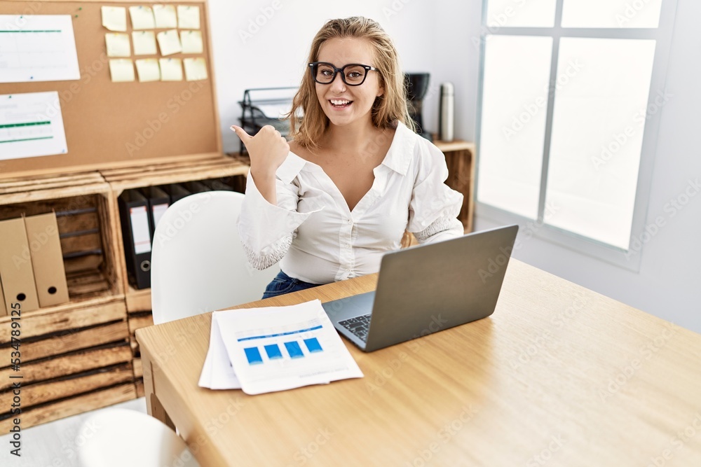 Poster young caucasian woman working at the office wearing glasses pointing thumb up to the side smiling ha