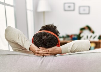 Young hispanic man on back view using headphones sitting on the sofa with hands on head at home.