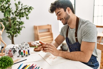 Young hispanic artist man smiling happy modeling pottery at art studio.