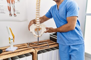 Young hispanic man wearing physio therapist uniform holding anatomical model of vertebral column at...