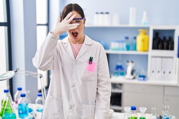 Young brunette woman working at scientist laboratory peeking in shock covering face and eyes with hand, looking through fingers afraid