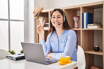 Young brunette woman wearing call center agent headset smiling with happy face winking at the camera doing victory sign. number two.