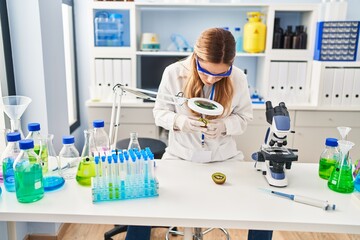 Young blonde woman wearing scientist uniform examining kiwi using magnifying glass at laboratory