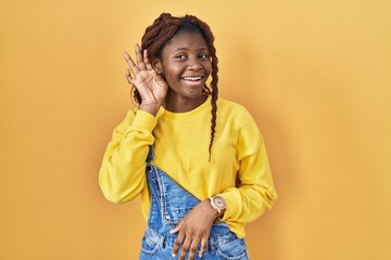 African woman standing over yellow background smiling with hand over ear listening an hearing to rumor or gossip. deafness concept.