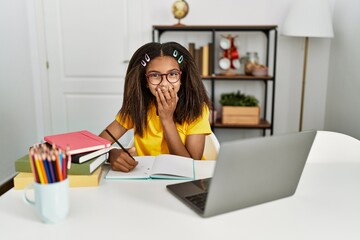 Young african american girl doing homework at home laughing and embarrassed giggle covering mouth...