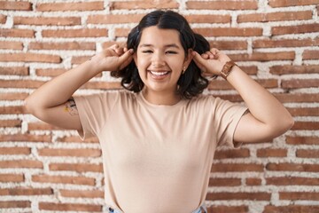 Young hispanic woman standing over bricks wall smiling pulling ears with fingers, funny gesture....