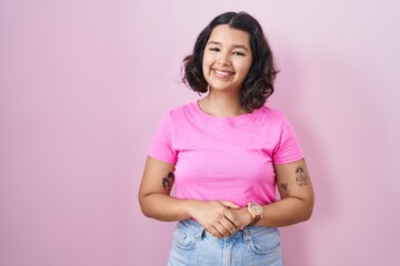 Young hispanic woman standing over pink background with hands together and crossed fingers smiling...