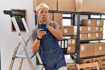Young blond man using smartphone working at storehouse bored yawning tired covering mouth with hand. restless and sleepiness.