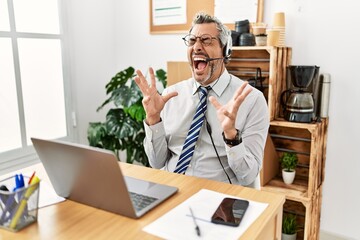 Middle age hispanic business man working at the office wearing operator headset celebrating mad and crazy for success with arms raised and closed eyes screaming excited. winner concept