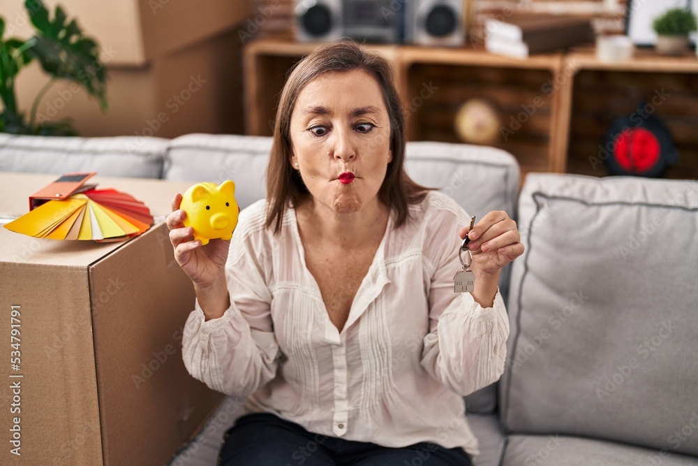 Sticker middle age hispanic woman holding piggy bank at new home making fish face with mouth and squinting e