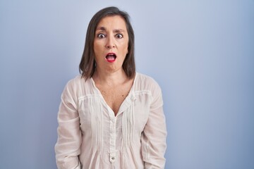 Obraz na płótnie Canvas Middle age hispanic woman standing over blue background in shock face, looking skeptical and sarcastic, surprised with open mouth