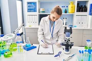 Young blonde woman wearing scientist uniform writing on checklist at laboratory