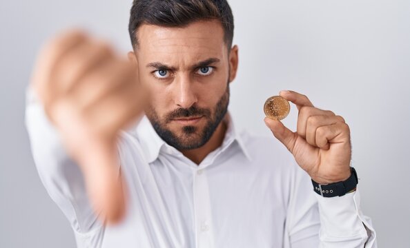 Handsome hispanic man holding cardano cryptocurrency coin with angry face, negative sign showing dislike with thumbs down, rejection concept