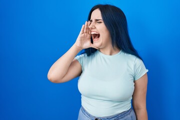 Young modern girl with blue hair standing over blue background shouting and screaming loud to side with hand on mouth. communication concept.