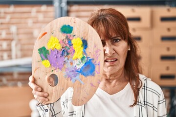 Middle age hispanic woman holding palette covering face clueless and confused expression. doubt...