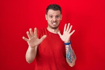 Fototapeta premium Young hispanic man standing over red background afraid and terrified with fear expression stop gesture with hands, shouting in shock. panic concept.