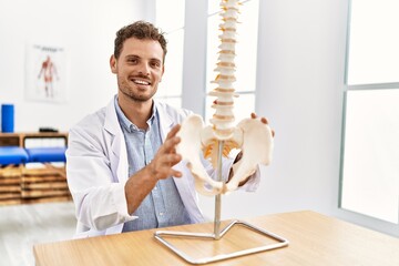 Young hispanic man wearing physiotherapist uniform touching anatomical model of vertebral column at...