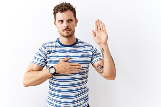 Handsome young man standing over isolated background swearing with hand on chest and open palm, making a loyalty promise oath