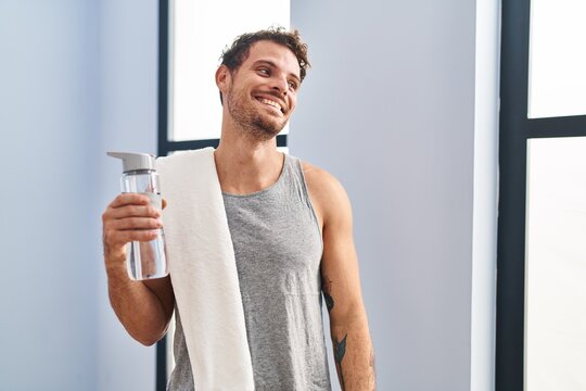 Young hispanic man wearing sportswear drinking water looking away to side with smile on face, natural expression. laughing confident.